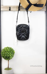 Black crossbody bag hanging on a wooden rack with a straw hat and potted plant below.