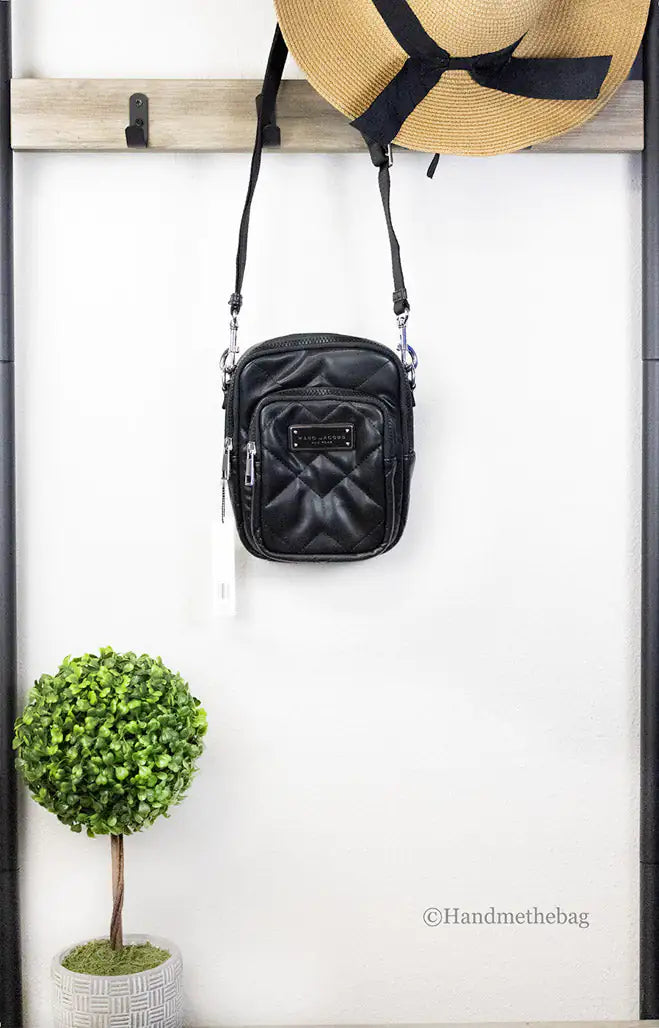 Black crossbody bag hanging on a wooden rack with a straw hat and potted plant below.